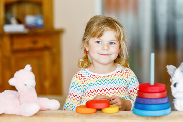 Cute little toddler girl playing alone with colorful wooden rainbow pyramid and toys at home or nursery. Happy healthy child having fun in kindergarten or preschool daycare