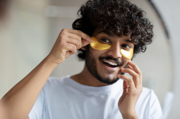 Skin care to reduce eye bags. Cheerful indian man applying gold anti-fatigue patches under eyes, looking at mirror