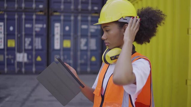 Black women engineer wears helmet and headphones Working at the container yard. She uses tablets to view container information waiting to be exported or imported in transportation logistic concept