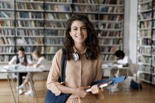 Happy Hispanic gen Z student girl with headphones visiting public library for work on study research project, holding learning papers, notebook, looking at camera, smiling. Head shot portrait