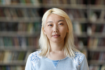 Pensive pretty Asian blonde student girl posing in university library with blurred background behind. Young Chinese hipster woman with pale hair looking at camera head shot portrait
