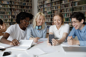 Happy cheerful multiethnic group of students sharing mobile phone, making video call in public college library, watching learning webinar, online lesson, consulting teacher, smiling