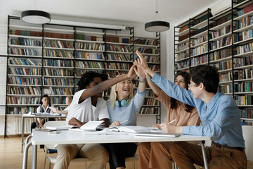 Interracial group of college girl and guys giving group high five gesture over table in university library, celebrating succes, completing project, laughing, smiling, expressing joy, unity