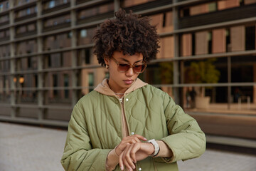 Stylish curly haired woman focused at watch checks time waits for someone at street wears stylish...