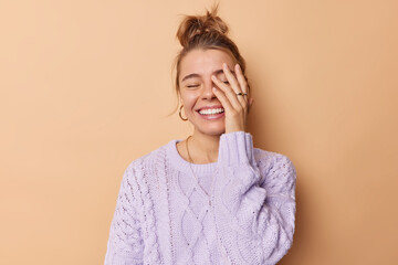 Happy carefree young woman makes face palm smiles broadly keeps eyes closed expresses positive emotions feels glad wears casual jumper isolated over beige background laughs at something funny