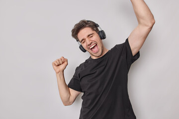 Overjoyed happy European man shakes arms exclaims from joy moves with rhythm of music listens music via wireless headphones wears casual black t shirt isolated over white background has fun.