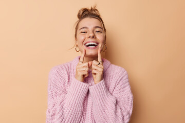 Positive young woman with combed hair points at her broad toothy smile feels very happy has white perfect teeth wears knitted sweater isolated over beige background. People and emotions concept - 482138178