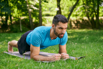 Naklejka na ściany i meble Athletic man doing plank exercise on yoga mat in park during sunny summer day