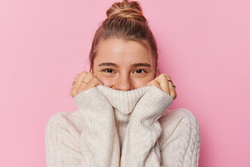 Positive young European woman with fair hair gathered in bun wears knitted warm white sweater during cold temperature weather covers mouth with collar looks happily isolated over pink background