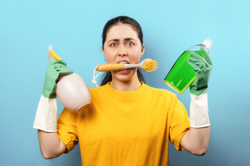 Portrait of a stressed Caucasian young woman in rubber gloves, holding a brush in her mouth and household cleaning products in her hands. Blue background. Cleaning day concept