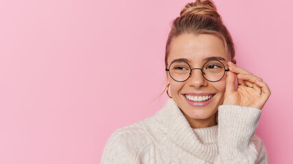 Portrait of cheerful young pretty woman keeps hand on rim of spectacles smiles broadly hears good news focused away wears white sweater isolated over pink background blak copy space for promotion