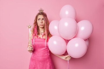 Obraz na płótnie Canvas Unhappy long haired young woman frowns face has bad mood holds magic wand bunch of inflated balloons wears crown and dress isolated over pink background. People holidays celebration concept.