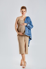 a beautiful pregnant blonde in a beige dress and jeans on one shoulder, standing on a gray studio background, pregnancy fashion
