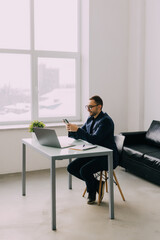 Business man sitting at desk in his office, holding phone and smiling happily while reading funny content on screen