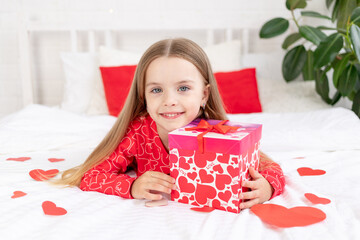 the concept of Valentine's day, a cute child girl is sitting on the bed at home in red pajamas and holding a gift in her hands and smiling or laughing with happiness, congratulating on the holiday