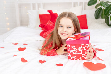 the concept of Valentine's day, a cute child girl is sitting on the bed at home in red pajamas and holding a gift in her hands and smiling or laughing with happiness, congratulating on the holiday