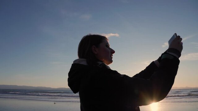 Young woman with cell phone, sunset beach, waves crashing, taking selfie video photo