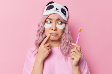 Thoughtful Asian woman with dyed hair looks attentively at toothbrush applies beauty patches under eyes has daily morning procedures wears sleepmask and casual t shirt isolated over pink background.