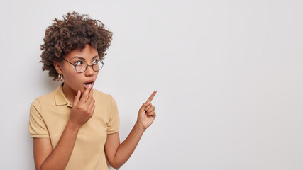 Studio portrait of amazed curly haired woman keeps jaw dropped from amazement stares bugged eyes...