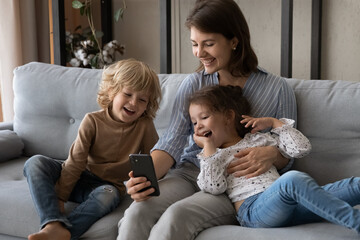 Joyful carefree little preschool kids siblings using smartphone with loving young mother, watching funny video or cartoons online in social network, playing entertaining mobile games together at home.