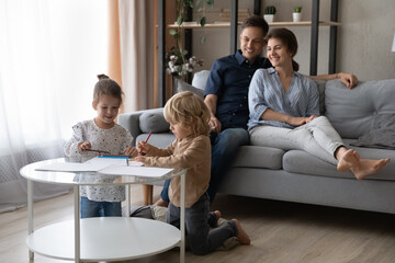 Happy millennial couple parents sitting on couch, watching small cute children siblings drawing pictures with colored pencils in paper album sitting at coffee table, friendly family leisure pastime.