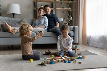 Happy friendly adorable little preschool kids boy girl playing favorite toys, sitting on floor carpet while caring smiling couple parents resting on comfortable couch at home, weekend relaxation time.