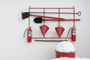 Pictured is the fire safety corner. White and red colors predominate. In the photo there are two shovels, fire extinguishers, cones, crowbars, hooks, a red barrel of sand covered with snow