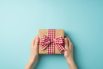 First person top view photo of valentine's day decorations female hands holding kraft paper giftbox with checkered ribbon bow on isolated pastel blue background with empty space