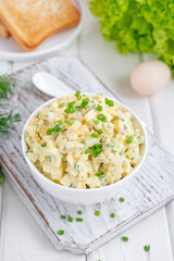 Egg salad with chopped green onions on top in a white bowl for cooking a sandwich on a white wooden background. Selective focus