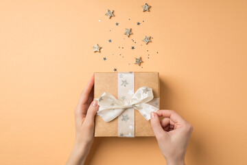 First person top view photo of valentine's day decor female hands untying white bow on craft paper giftbox glowing stars and star shaped confetti on isolated beige background
