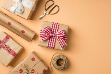 Top view photo of st valentine's day decor craft paper gift boxes with bows scissors spool of twine and roll of kraft paper with polka dot pattern on isolated beige background with copyspace