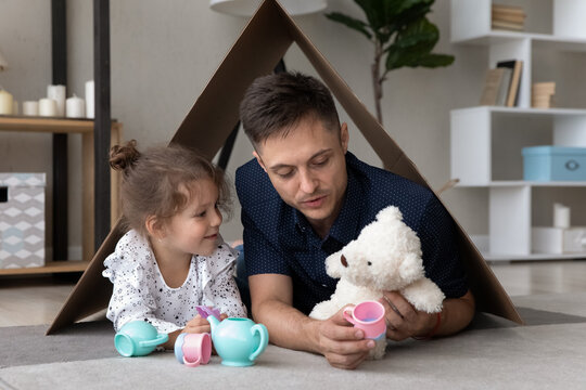 Happy little cute kid girl lying with caring single dad on floor carpet under carton roof, playing entertaining game together at home, imagining playing toy tea ceremony, family weekend pastime.