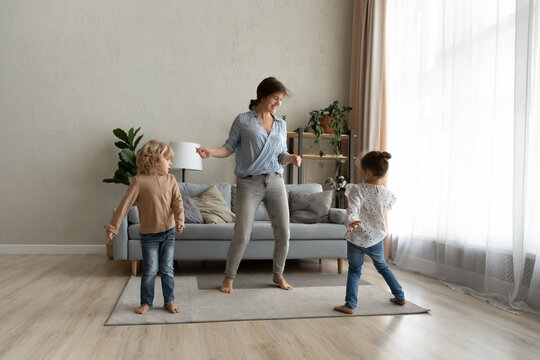 Laughing happy young single mother dancing barefoot on floor carpet with joyful little kids son daughter, listening energetic disco pop music together at home, family hobby leisure activity concept.