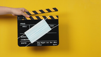 Naklejka premium The hand is holding a black clapper board with a medical face mask on yellow background.