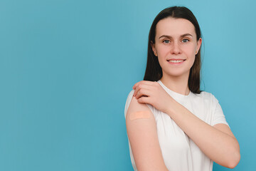 Obraz premium Portrait of smiling cute young caucasian female after getting vaccine, posing isolated on blue wall. Woman holding down her t-shirt sleeve and showing her arm with bandage after receiving vaccination