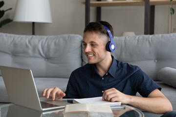Smiling young man in wireless headphones web surfing educational information on laptop, improving...
