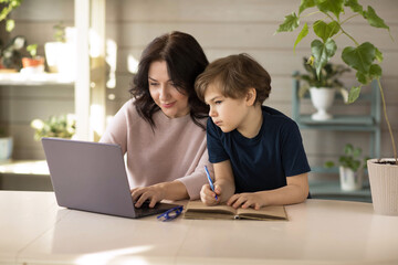 Mother and son are engaged in online school, sit in a laptop and make notes in a notebook