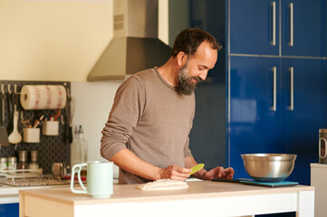 Smiley bearded adult male looking at his smart phone while trying to make some bread at home