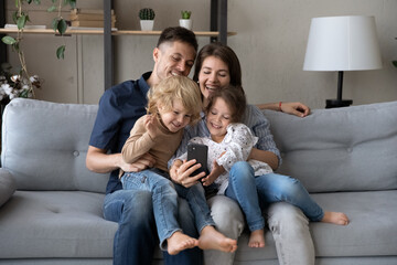 Happy bonding young mother and father holding on laps small adorable kids son and daughter, having fun playing games on cellphone, recording selfie video, posing for photo or enjoying web camera call.