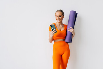 Young athletic woman holding rubber mat for yoga and using mobile phone, standing against white...