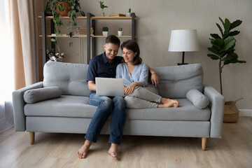 Obraz premium Joyful bonding young family couple using laptop, resting on comfortable couch, enjoying web surfing information, booking flight tickets or hotel online planning trip, shopping in internet store.