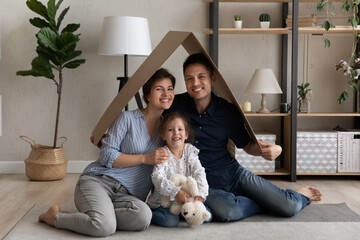 Portrait of loving caring young couple parents and small cute kid daughter sitting in modern renovated living room under cardboard roof, feeling excited purchasing own dwelling, real estate concept.