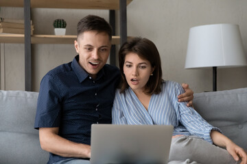 Fototapeta premium Surprised young bonding family couple looking at computer screen, getting email with amazing unbelievable news, reading message with online lottery gambling auction win notification, internet success.
