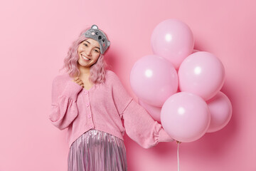 Obraz na płótnie Canvas Positive carefree woman with eastern appearance has glad expression dressed in jumper and pleated skirt has happy festive mood poses with balloons isolated over pink background. Celebration concept