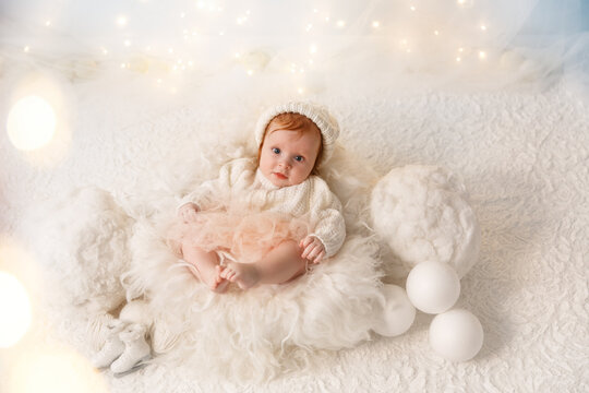 Little red hair baby on the white wool. Infant girl wearing white sweater and hat in photo studio which makes like winter composition. Garlands are behind the child.