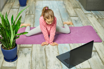 Little girl watching online video on laptop and doing stretching, fitness exercises at home