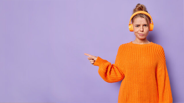 Horizontal Shot Of Disappointed Young European Woman With Combed Hair Frowns Face Feels Unhappy Wears Stereo Headphones On Ears Shows Blank Copy Space Against Purple Backgrouund. I Dont Like It