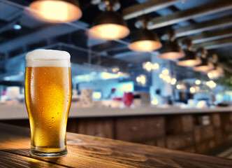 Cooled glass of beer on the wooden table. Blurred bar interior at the background.