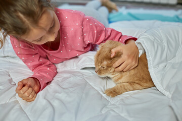 Cute ginger cat and little girl lying on a soft blanket in bed