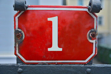 A red house number plaque, showing the one (1)